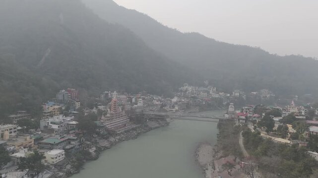 Aerial view of Lakshman Jhula in Rishikesh, Uttarakhand, with the Ganga River flowing below, and temples, houses, and buildings along the riverbank against misty mountains on a winter morning.