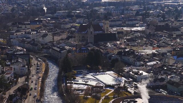 Aerial view of Brixen, Italy shows the duomo and clock tower, the Isarco River, cars on the riverside road, steam from a modern rooftop, and a frozen pool with slides.
