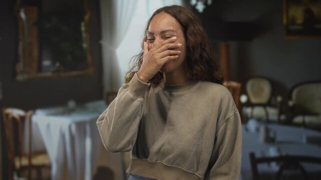 Woman hand covering mouth laughing in restaurant dining room, casual sweatshirt and uncovered hand visible; amusement connection.