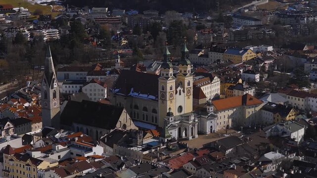 Aerial pan and descent reveal Brixen Cathedral twin domes and Gothic spire, clock faces, Baroque facade, snow patches, red tiled blocks, long winter shadows.