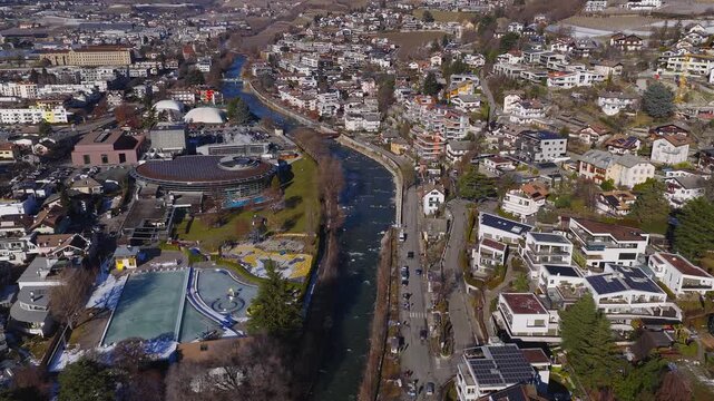 Aerial view of Brixen Bressanone, South Tyrol, Italy, shows Eisack River, small bridges, riverside road, leisure complex with solar roof, cars, steam, bare trees, snow patches.