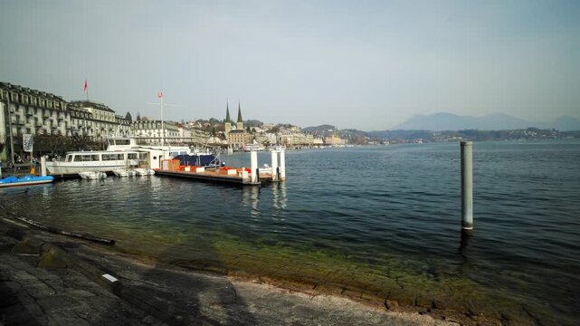 Static wide shot of the Lake Lucerne waterfront featuring a boat pier, historic architecture, and the twin towers of Hofkirche St. Leodegar