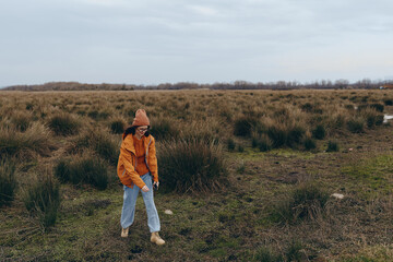 Naklejka premium Woman lifestyle concept with a young woman wearing an orange jacket and hat walking in a vast grassy field under an overcast sky during autumn season outdoors.