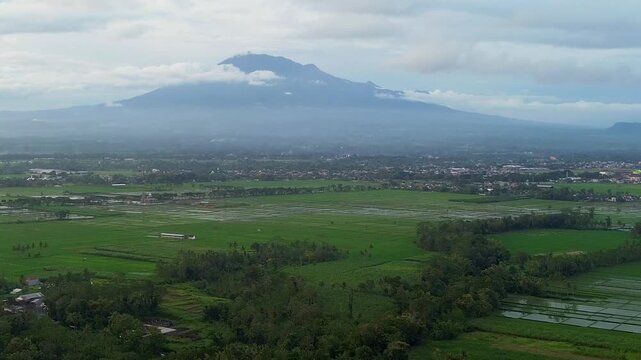 Aerial panoramic view of Kawi and Arjuno mountains peak over lush green rice fields in East Java.