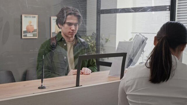 Young male customer holding documents, interacting with bank teller at counter, separated by transparent protective screen