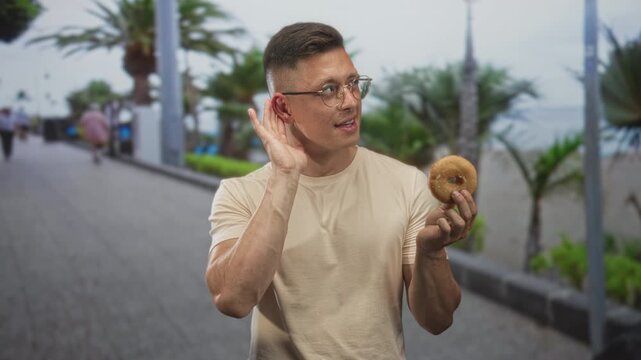 Man holding donut and cupping ear gesture on street promenade by palm trees and ocean; playful curiosity.