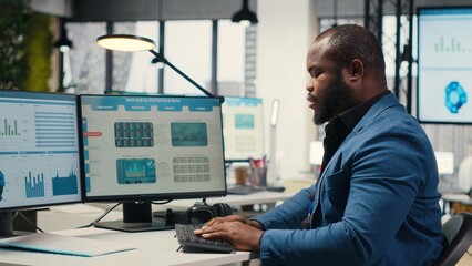 Black male analyst focused on IT company development with digital tools, review analytics and performance metrics at workstation. Staff planning solutions and aligning objectives. Camera B. © DC Studio