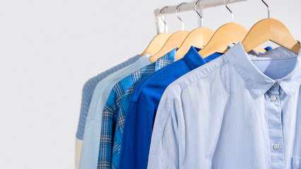Fototapeta premium Close up blue shirts hanging on wooden beige hangers on clothing rack, light gray background. Concept of capsule wardrobe, minimalist clothing basics and organized closet lifestyle. Copy space, banner