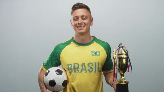 Young man wearing yellow brazil jersey with flag patch and brasil text holds soccer ball in left hand and gold trophy in right, smiling pose and chest forward in studio; pride.