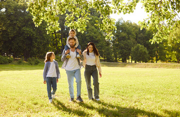 Fototapeta premium Happy family with two kids walking together in sunny park. Smiling father carrying daughter on shoulders while mother and son strolling beside. Parents enjoying outdoor leisure time with children.