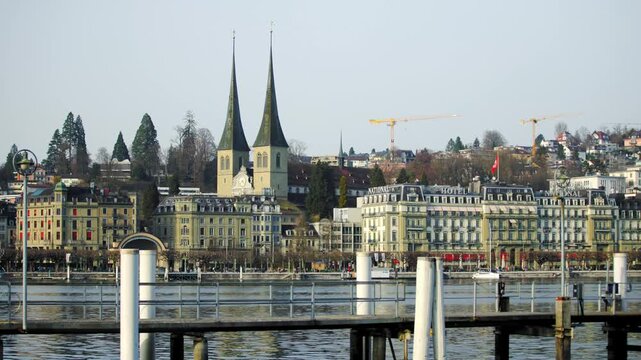 LUCERNE, SWITZERLAND - MARCH 06 2026: Static wide shot of the Lake Lucerne waterfront featuring the historic Hofkirche St. Leodegar and the Grand Hotel National on a clear day.