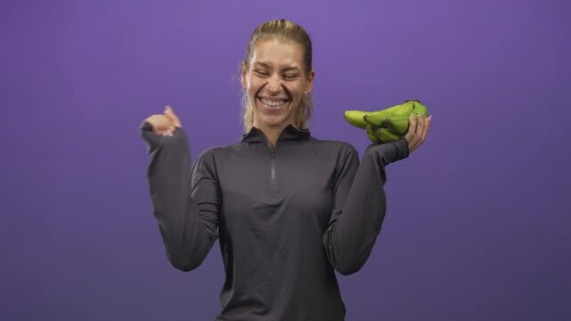 Woman holding a bunch of green bananas in one hand while waving with the other hand in a studio, smiling and wearing a dark zippered pullover; cheerful healthy snack.