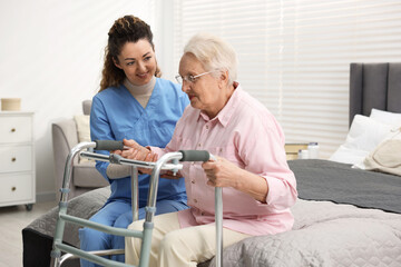Obraz premium Care worker helping senior woman with walker to stand up from bed indoors