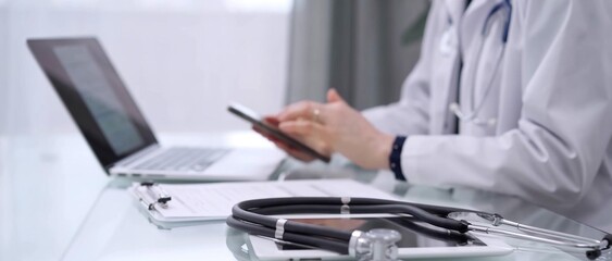 Fototapeta premium Stethoscope and clipboard are lying on the glass table while doctor woman is using a smartphone at the background. Medicine concept