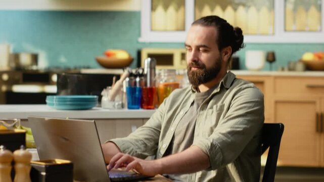 Freelancer in casual attire working remotely at kitchen table with laptop. Web browsing and connection support productivity in modern home setup, showcasing flexible work and technology use.