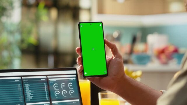 Vertical chroma key screen on smartphone used at home in kitchen, remote worker analyzing metrics and charts on laptop. Teleworking and outsourcing showing task management and research.
