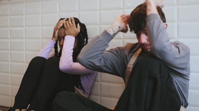 Two tired multiracial baristas colleagues sitting inside a cafe with their hands on their heads while facing a difficult moment during work hours in the afternoon