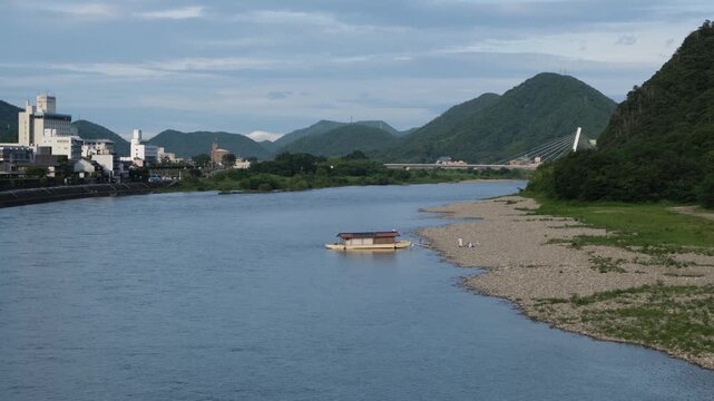 Scenic view of the Nagara River in Gifu City, Japan, with calm flowing water, surrounding mountains, and low-rise urban buildings in town along the riverbank. A peaceful regional city. Slow living.