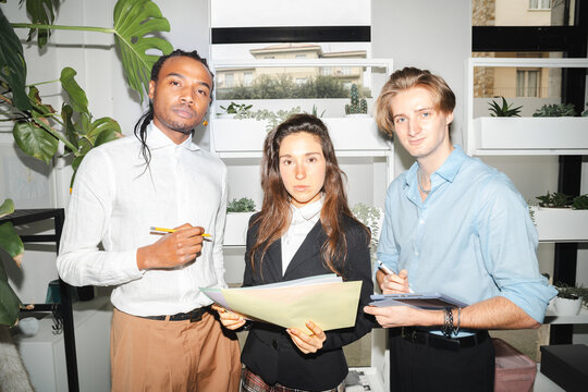Three young professionals, two men and one woman, collaborate on a project, reviewing documents and taking notes in a modern office setting with plants.