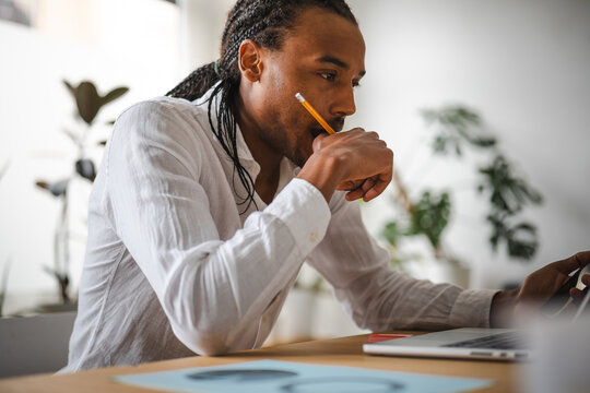 A man with braided hair holds a pencil to his chin while looking at a laptop screen, deep in thought.