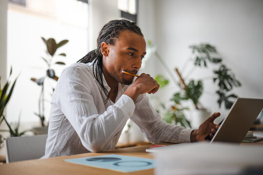 A man with braided hair, wearing a white shirt, sits at a desk with a laptop and a pencil in his mouth, deep in thought.