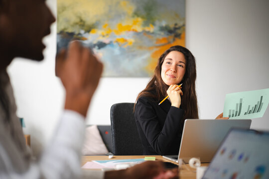 A woman in a black blazer smiles while holding a bar graph and a pencil, sitting at a desk with laptops and papers.