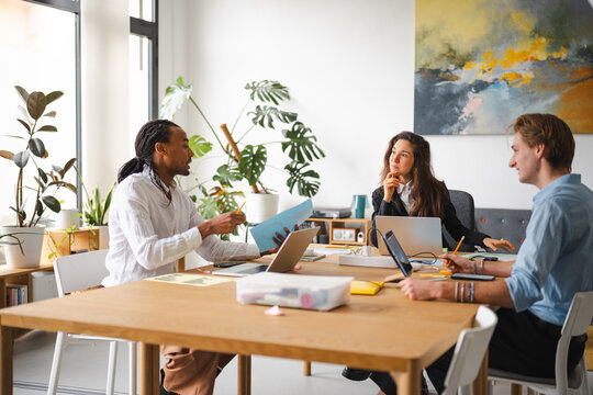 A diverse team collaborates around a table, discussing documents and using laptops in a bright, modern office setting with plants.