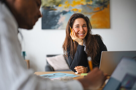A woman smiles while working with a colleague in an office setting, with charts and a laptop on the table.