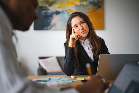 A woman in a black blazer and white shirt sits at a desk, holding a pencil to her chin, looking thoughtfully at the camera while a colleague works on a laptop.