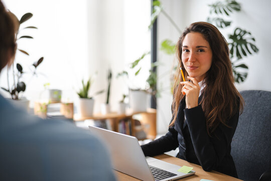 A woman in a suit holds a pencil to her chin while looking at a laptop. She is in an office setting with plants in the background.