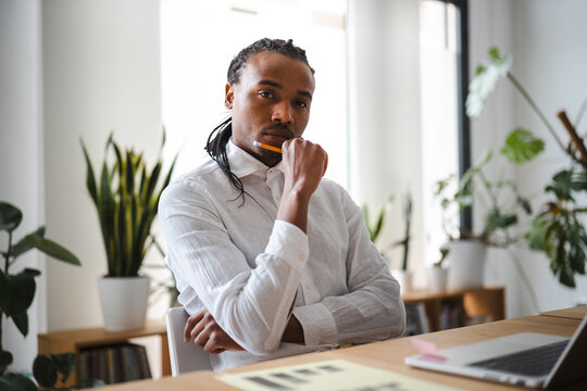 A man with dreadlocks sits at a desk, holding a pencil to his chin, deep in thought. He wears a white linen shirt and is surrounded by plants.