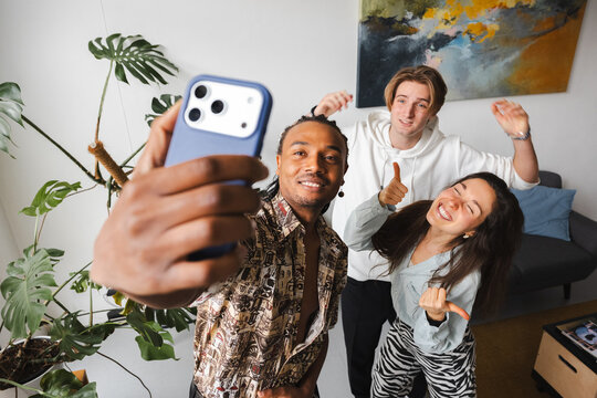 Three friends pose for a selfie in a bright, modern living room. One person holds the phone, while the other two make fun gestures.