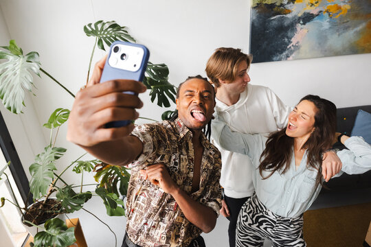 A man takes a selfie with his friends, sticking his tongue out and winking. His friends laugh and pose behind him, enjoying a fun moment together.