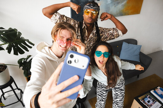 Three friends pose for a selfie, two wearing heart-shaped sunglasses and one flexing his muscles.