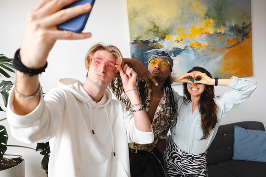 Three friends pose for a selfie, two wearing heart-shaped glasses and one making a heart shape with their hands.