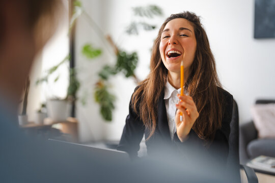 A woman in a suit laughs heartily while holding a pencil, engaged in a conversation with someone across a table in a bright, modern office setting.