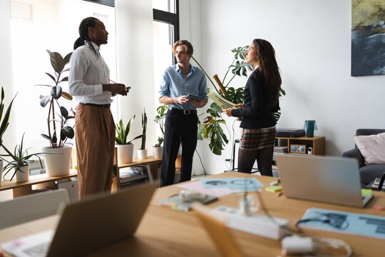 Three professionals discuss a project in a bright, modern office space with plants and laptops on a table.