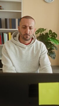 Man at computer with hand on chin and furrowed brow, wearing hoodie, looking at monitor with sticky note and bookshelf behind in a building; concern.