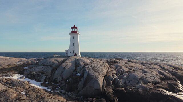 Drone aerial shot: Peggy's Cove lighthouse and snowy granite rocks at sunset over Atlantic Ocean. Halifax suburb Nova Scotia Canada, dramatic winter seascape with golden light 4K.