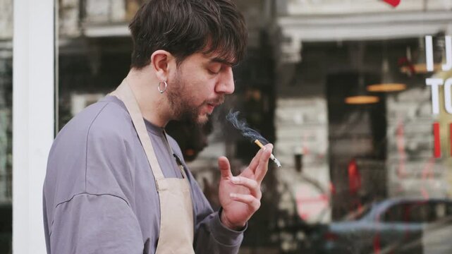 Barista on break outside cafe with cigarette while colleagues serve customer in busy urban area, smoking and using smartphone