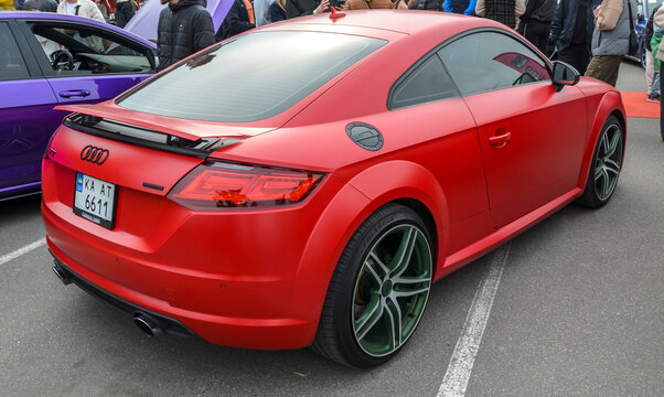 Matte red Audi TT with a black grille and matching mirrors sits at an outdoor event. It features sleek LED headlights. A smooth, modern look for a sporty coupe