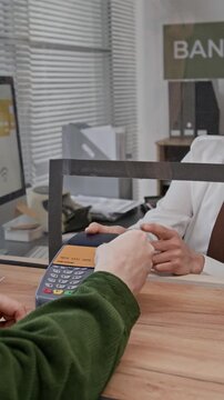 Vertical shot of focused young man entering pin on card machine, teller observing transaction across transparent divider