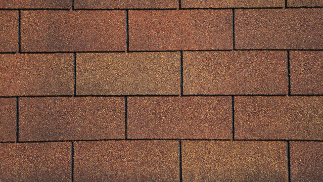 A close-up of brown and tan asphalt roof shingles arranged in a staggered rectangular pattern. The surface features a gritty, granular texture with subtle variations in earth-toned shades.