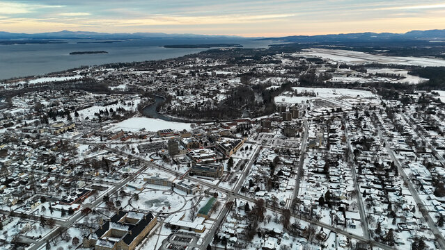 Plattsburgh, new york cityscape covered in snow, showcasing the saranac river winding through the urban area and lake champlain extending to the mountains under a winter sky