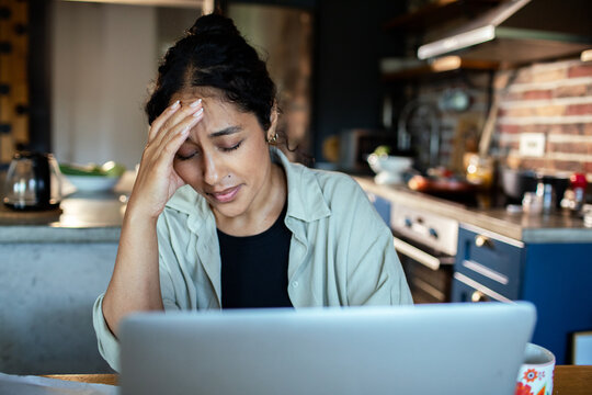 Stressed woman with headache working on laptop at home kitchen