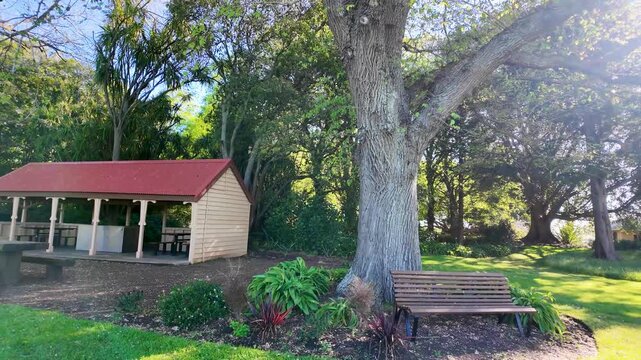 Shaded seating in Camperdown Botanic Gardens, VIC Australia, featuring a classic public pavilion and wooden bench under lush trees. A peaceful park and a regional attraction for recreational activity