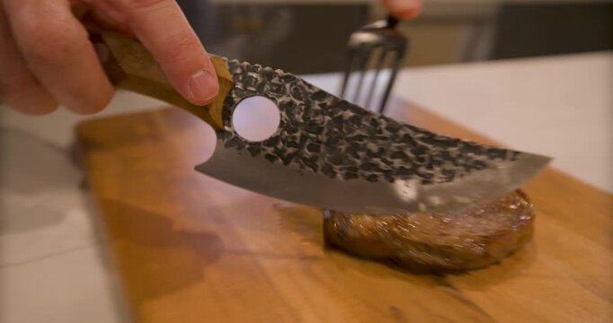 Closeup of fork moving sizzling steak to wooden cutting board and using sharp knife to slice it