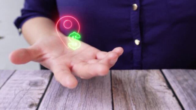 Geographic value. Close-up of a woman's hand on a table holding a virtual location pin icon with a dollar sign.