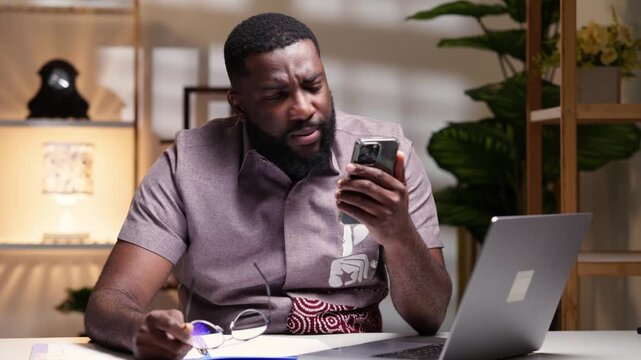African man receiving shocking news on a smartphone while sitting at an office desk