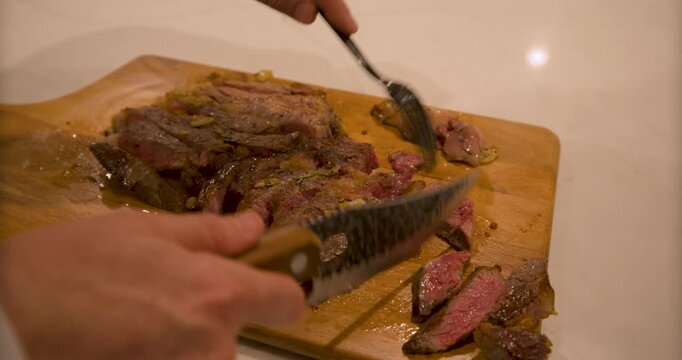 Cook with fork and knife cutting steak into smaller pieces on wooden board - closeup